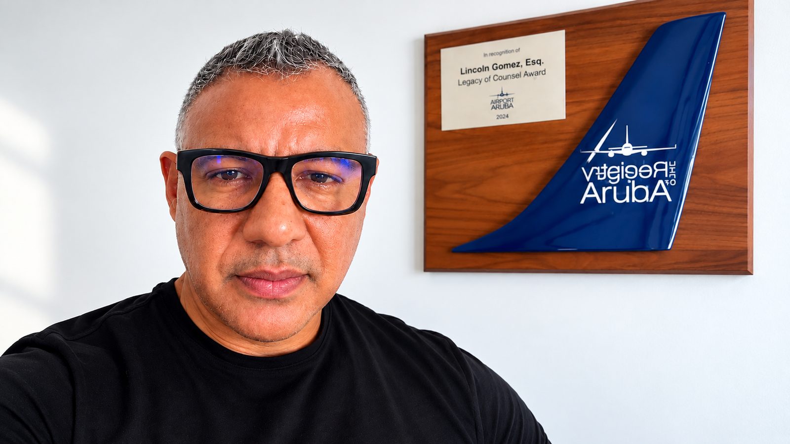 Portret van mr. Lincoln D. Gomez in een zwart T-shirt, met op de achtergrond een plaquette met een blauwe vliegtuigstaart.