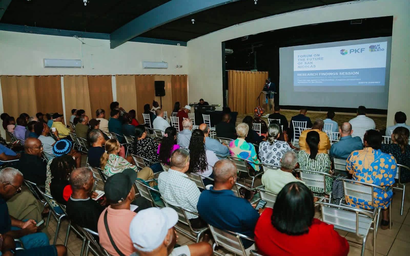 The audience at the Santa Teresita Center in San Nicolas, every seat taken, facing a large screen showing the title slide of the Forum on the Future of San Nicolas research findings session