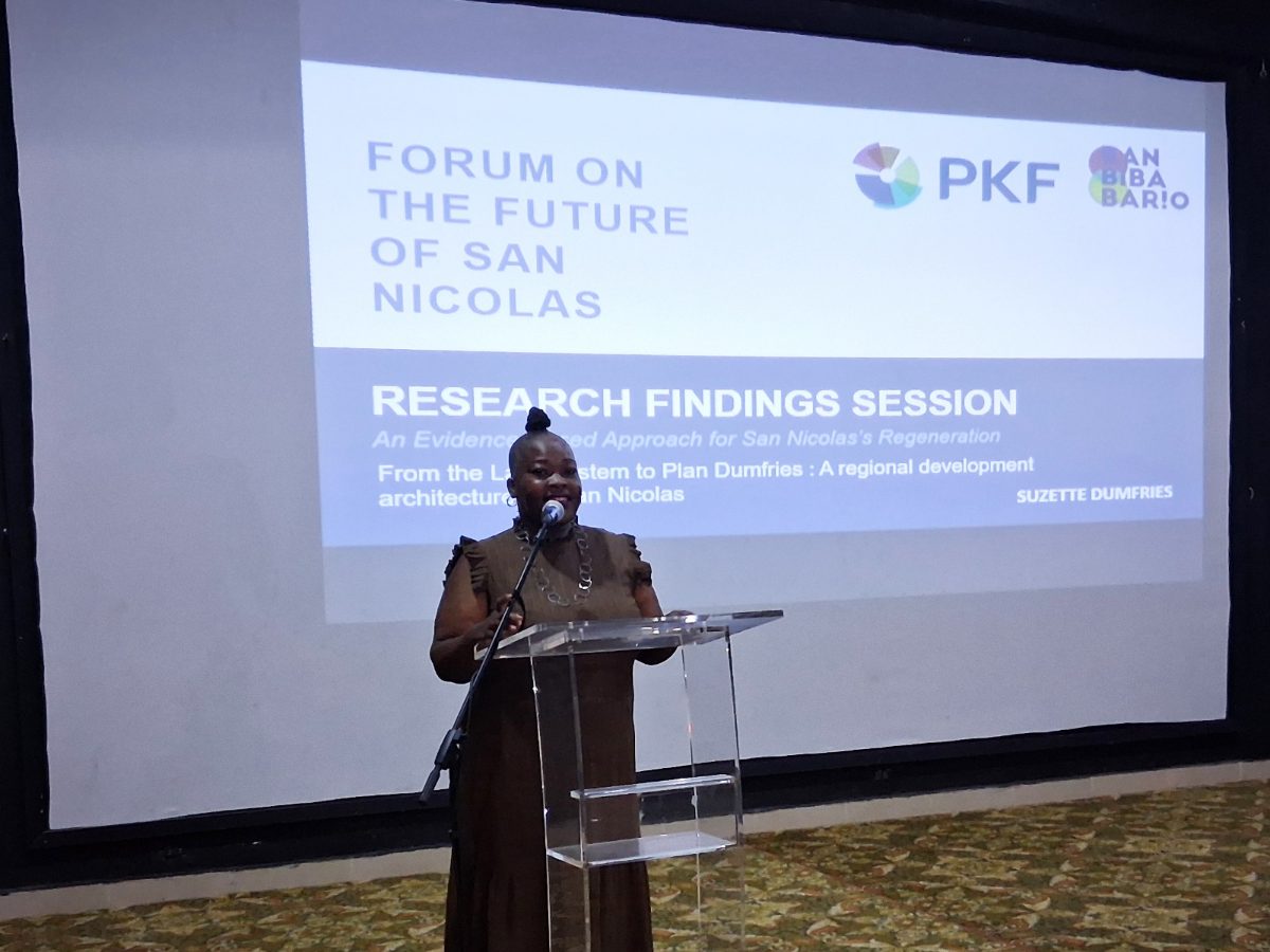 Suzette Dumfries speaking at a clear lectern in front of a projection screen reading Forum on the Future of San Nicolas, Research Findings Session