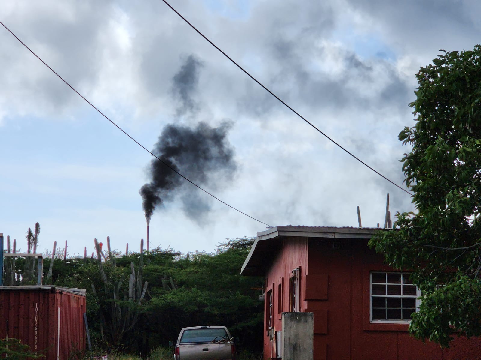 Chimney emitting black smoke beyond scrubland, viewed from a residential yard