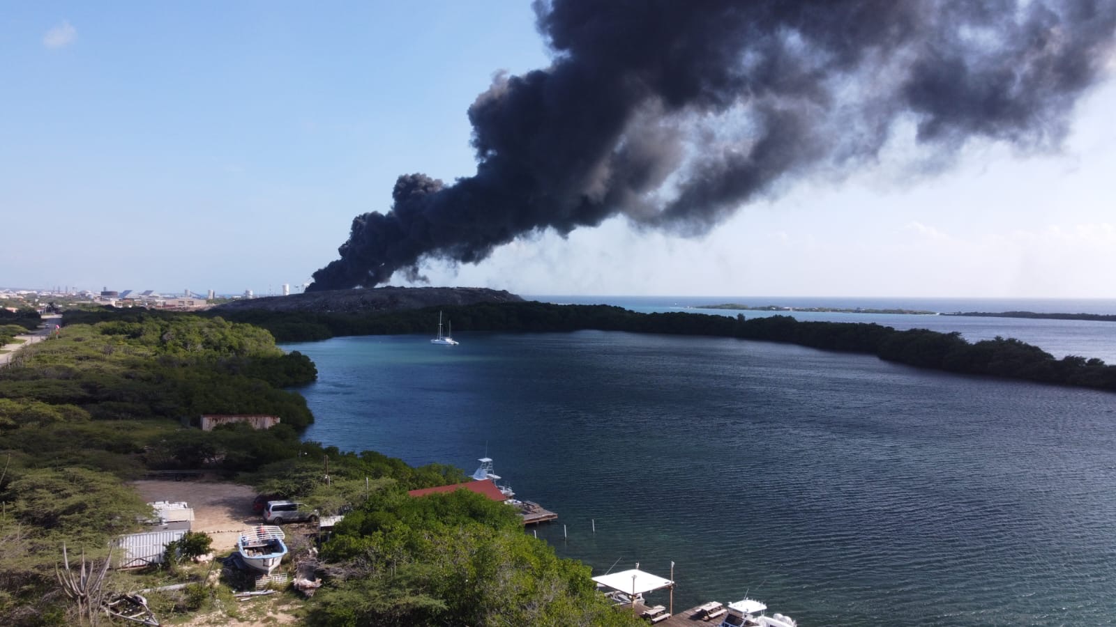 Aerial view of smoke drifting from a landfill across a mangrove lagoon with moored boats