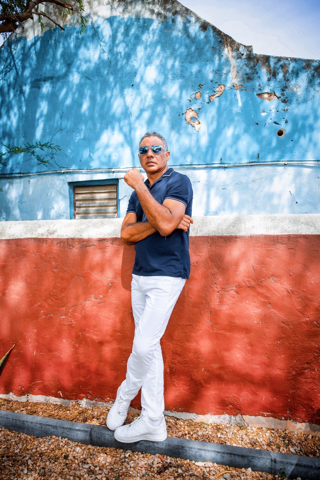 Man in a polo shirt and sunglasses leaning against a weathered blue and red wall