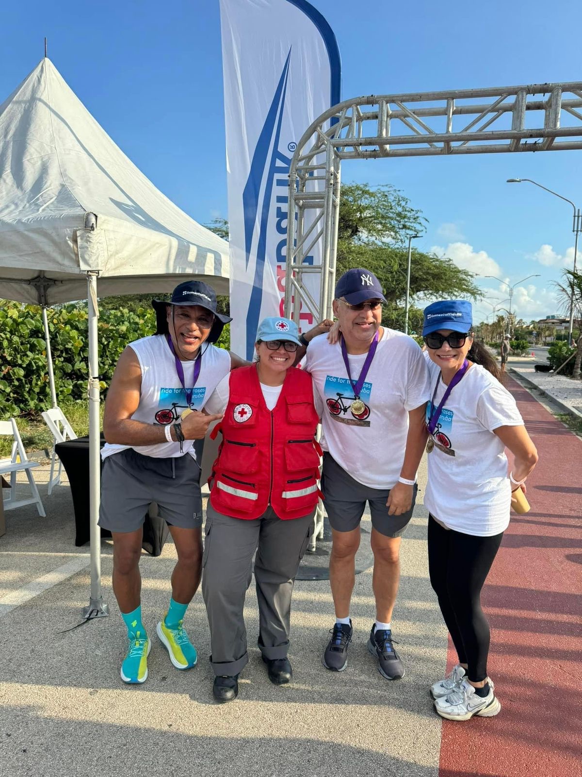 Four people, one in a Red Cross Aruba vest, posing together at a walk event finish arch