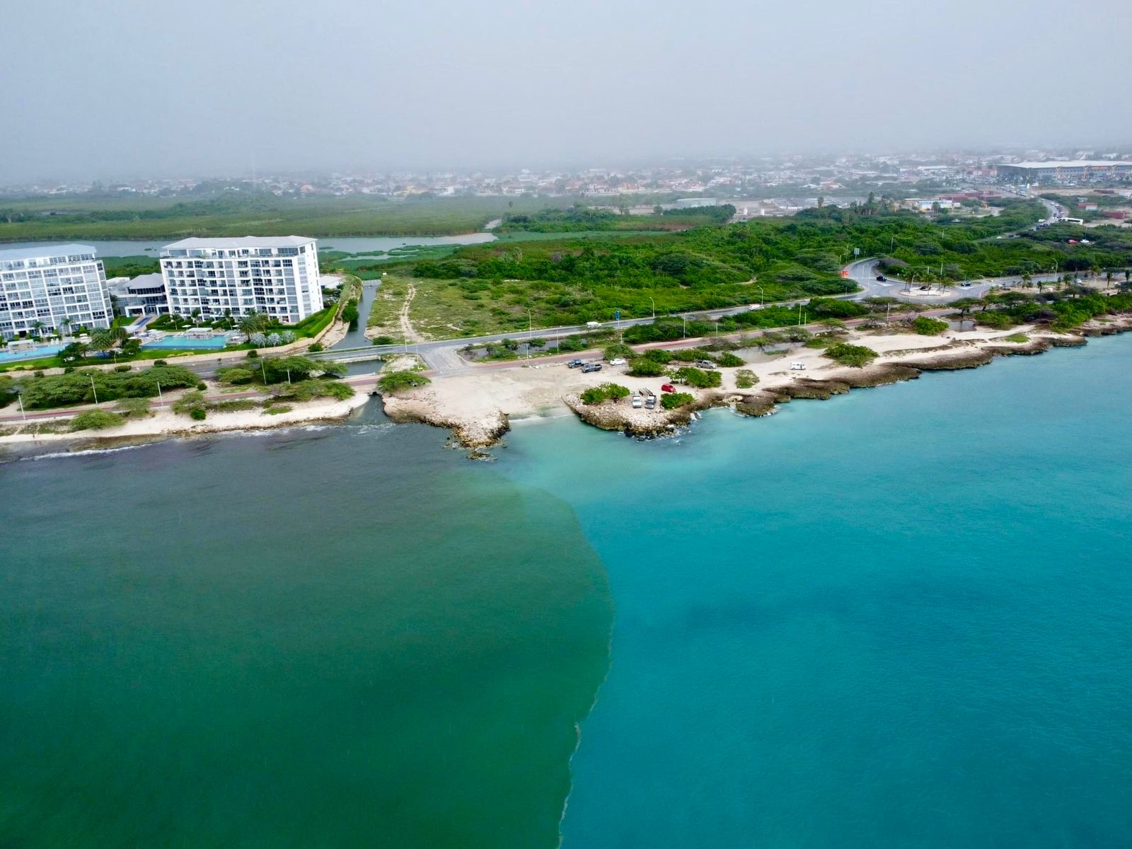 Aerial view of brown runoff discharging into turquoise coastal water beside apartment blocks
