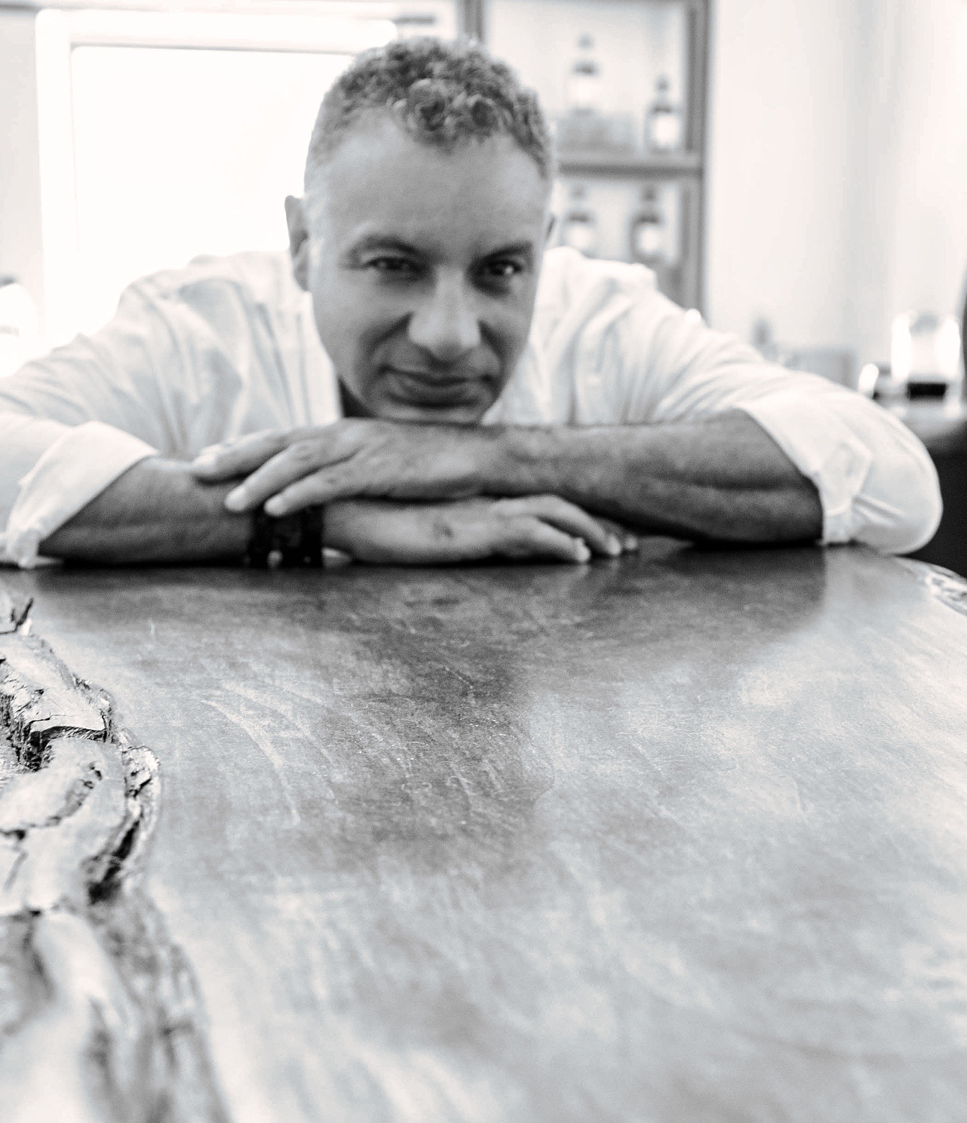 Black and white portrait of a man resting his chin on his folded arms at a wooden table