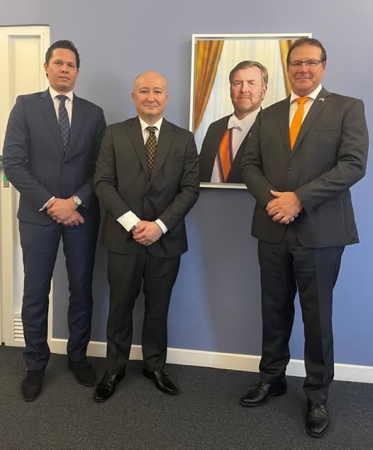 Three men in suits standing beside a framed portrait of the King of the Netherlands