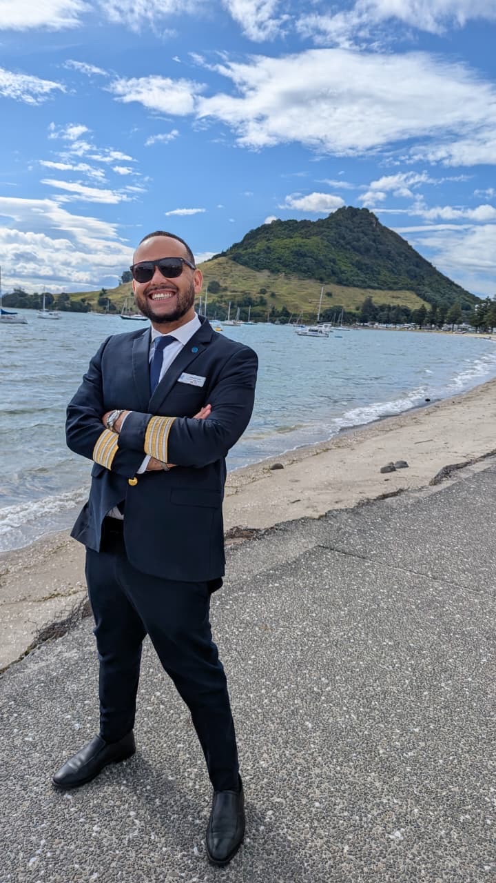 Man in a uniform blazer with gold sleeve braid, standing on a shore before a headland