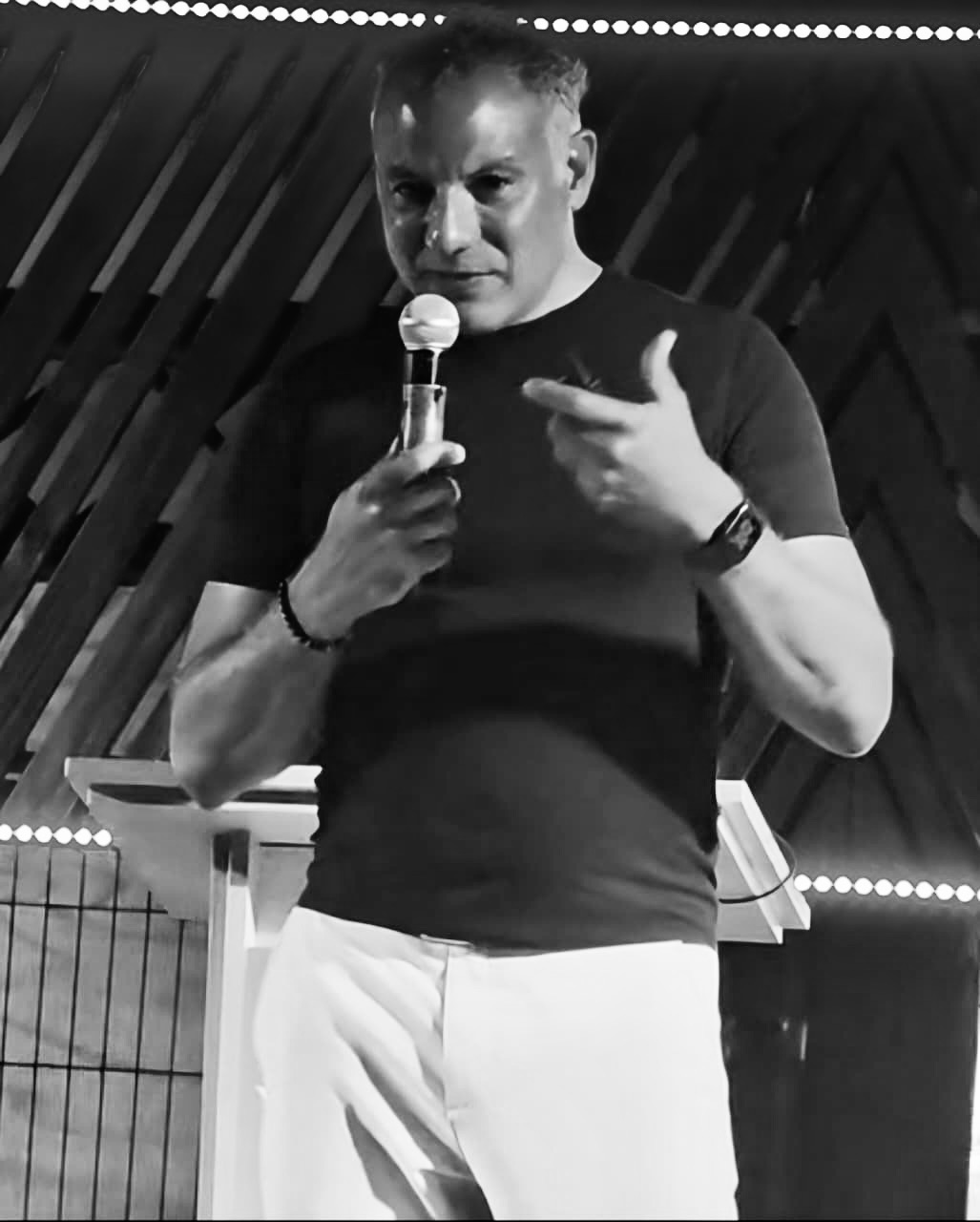 Man speaking into a handheld microphone on stage under strings of lights